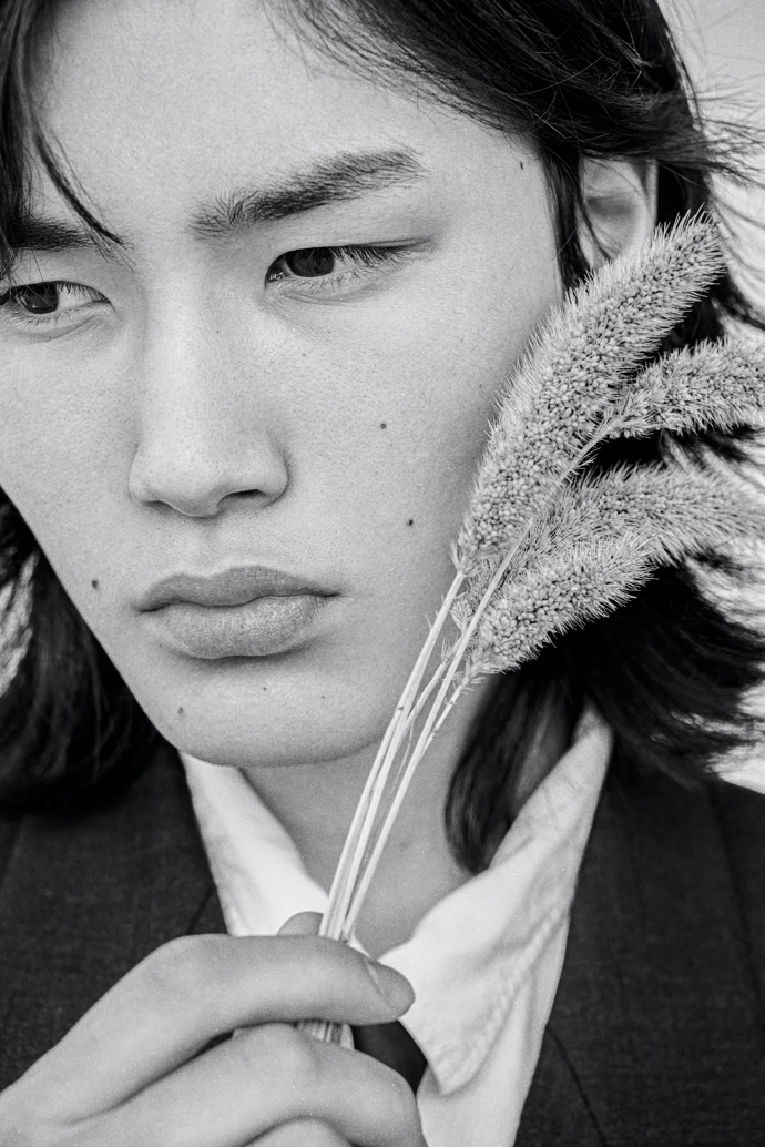 A moody greyscale professional photo of person in a suit and tie holding a feather-like grass against their face